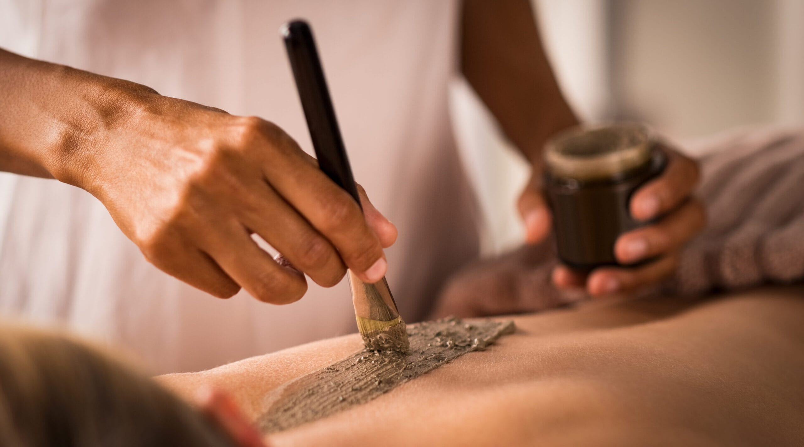 Closeup of beautician applying clay with brush on back of woman lying on massage table at spa. Woman hands using brush for clay mask. Beautiful lady receiving a scrub exfoliation therapy on body in a wellness center.