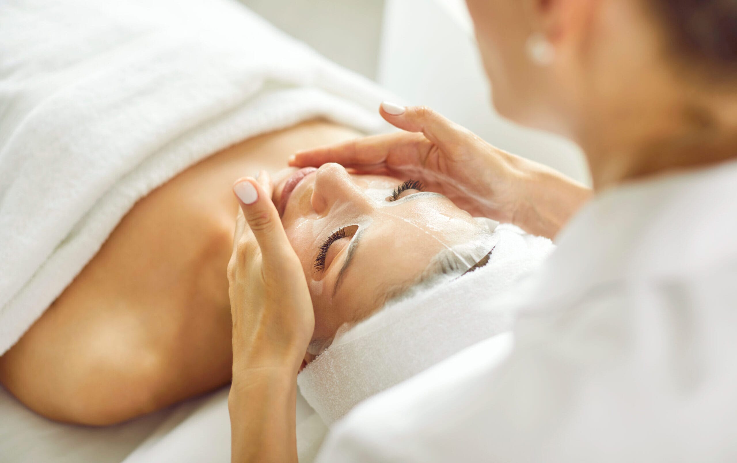 Professional cosmetologist at spa salon applying sheet mask on face of relaxed woman. Young girl lying on white towel and enjoying peeling and antioxidant facial beauty treatment. Skin care concept